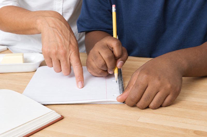 Un enfant écrit au crayon sur un cahier, guidé par une main adulte